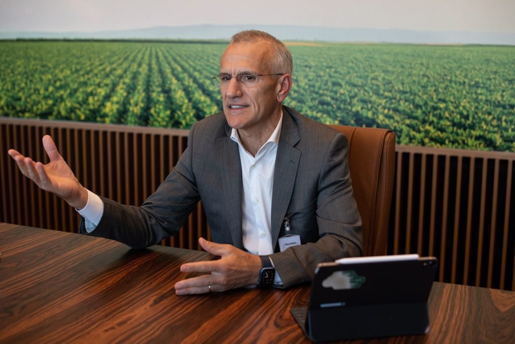 Homem de terno cinza sentado à mesa de madeira escura, gesticula. Ao fundo, grande imagem de um vasto campo agrícola verde.
