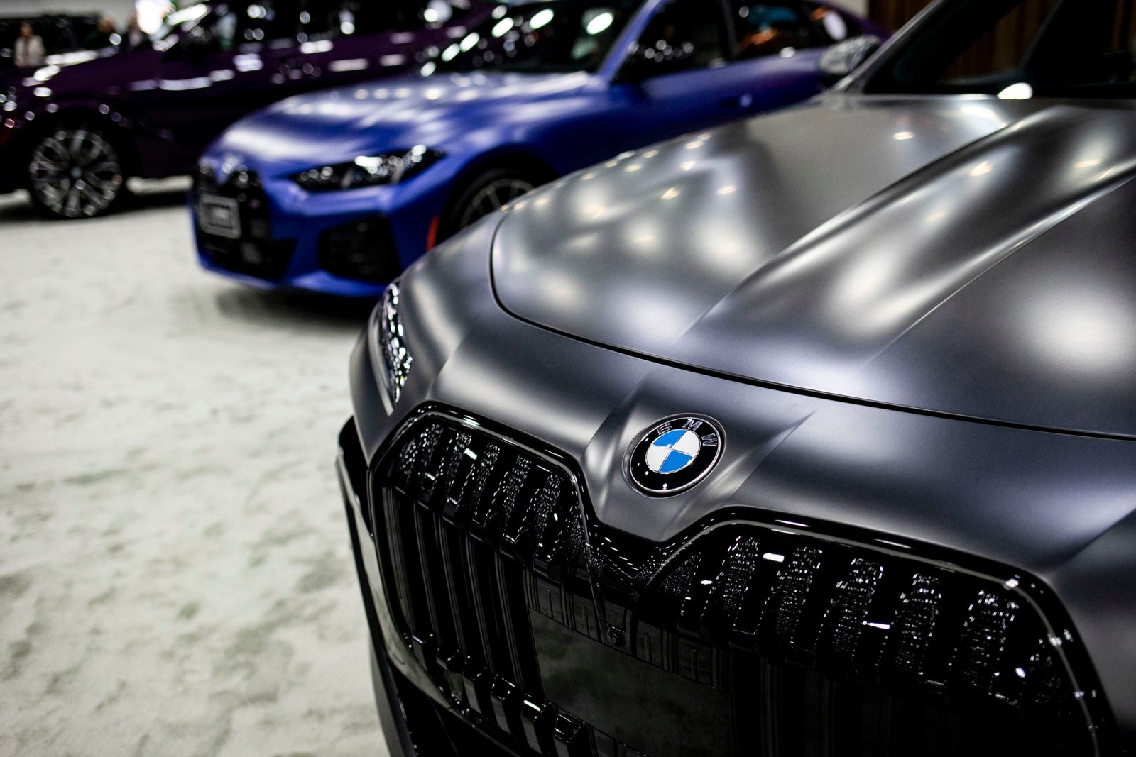 Frente de um BMW cinza fosco com logo em destaque. Dois carros desfocados, azul e roxo, estão ao fundo.